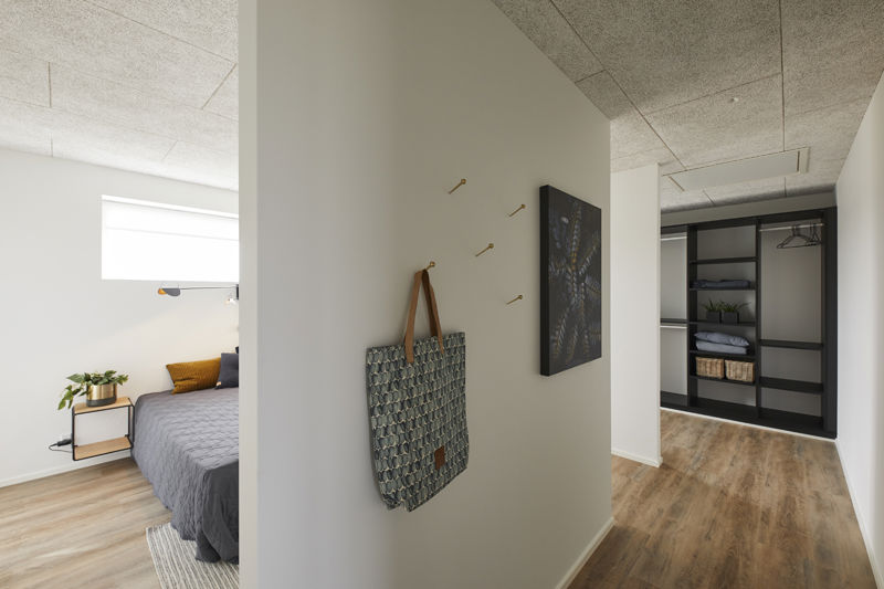 Soveværelsesgang fra eurodan-huse med vægkroge, mønstret taske, kunst på væggen, åbent walk-in closet med sorte reoler, kurve, hylder og bøjlestang samt kig til soveværelse – inspiration til praktisk opbevaring i nybyggeri og husbyggeri.