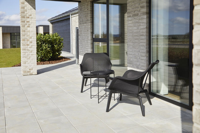 Overdækket terrasse fra eurodan-huse med lyse murstenssøjler, store gulv-til-loft vinduer, mørke loungestole, lille rundt bord og grå terrassefliser langs facaden – inspiration til nybyggeri med læ, udeliv og moderne terrassemiljø.