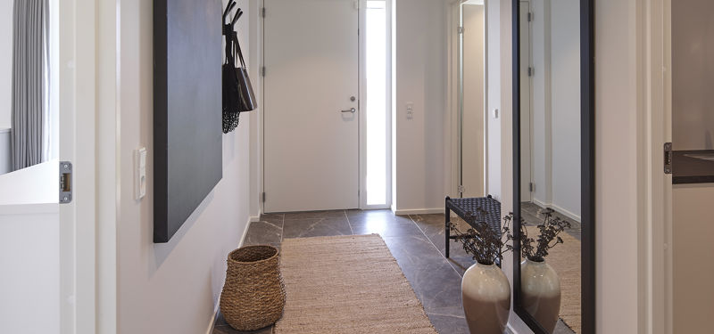 Lys entré med hoveddør og smalt sidevindue, mørke gulvfliser, lang løber, sort vægpanel med knager, stor spejlvæg og bænk – inspiration til en funktionel entré i nyt hus.