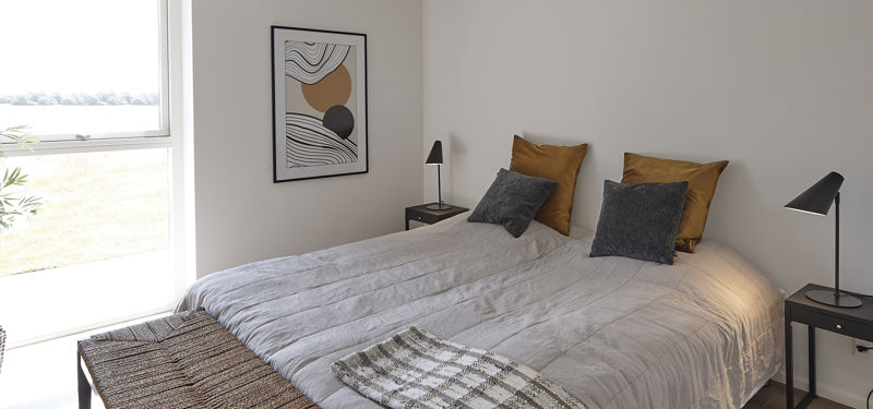 Lyst soveværelse med dobbeltseng, gråt sengetøj, sennepsgule puder, sorte væglamper, vævet bænk og stort vinduesparti – inspiration til indretning i et nyt hus.