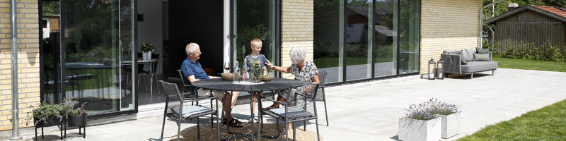 Tre personer nyder et måltid på den solrige terrasse ved et nyt eurodan-hus med store glaspartier.