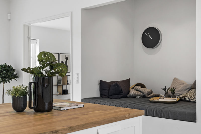 Indbygget siddealkove fra eurodan-huse med grå hynde, mørke puder, tæppe, lille bakke, rund vægur på væggen og køkkenø med træbordplade og grønne planter i forgrunden – inspiration til nybyggeri og husdrømme med praktisk hyggeplads.