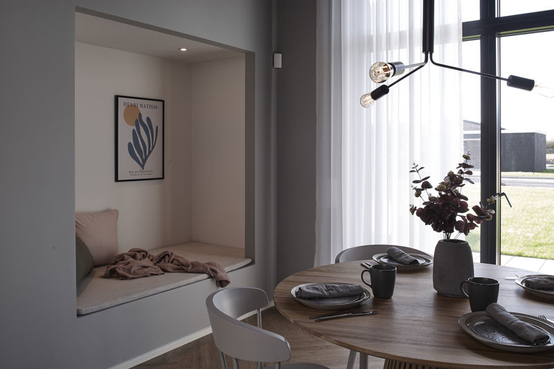 Rund spiseplads fra eurodan-huse med dækket træbord, store sorte vinduespartier, lette gardiner, moderne pendel og indbygget alkove med lys hynde, plaid og vægplakat – inspiration til nybyggeri og husdrømme med en hyggelig krog ved alrummet.