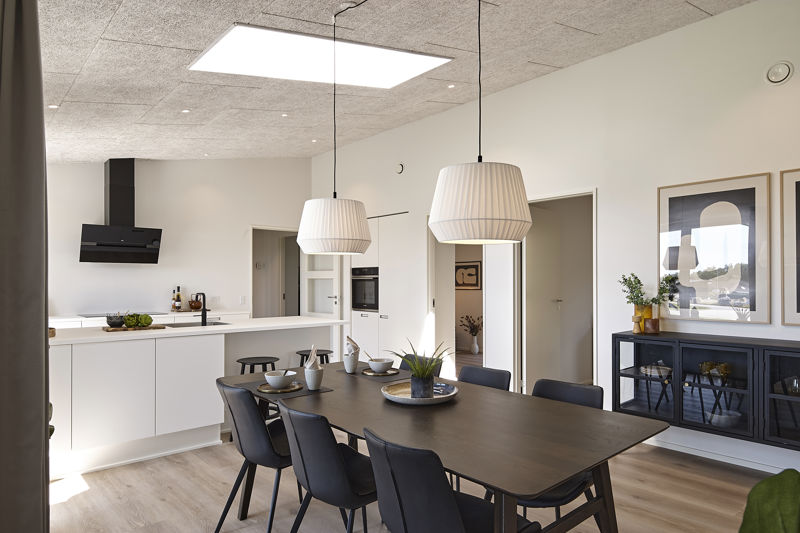 Spiseplads og køkken fra eurodan-huse med stort mørkt spisebord, sorte stole, to hvide pendler, sort vitrineskab med billeder over, hvid køkkenø, sort emhætte, lyse gulve og ovenlys i loftet – inspiration til åbent køkken-alrum i moderne nybyggeri.