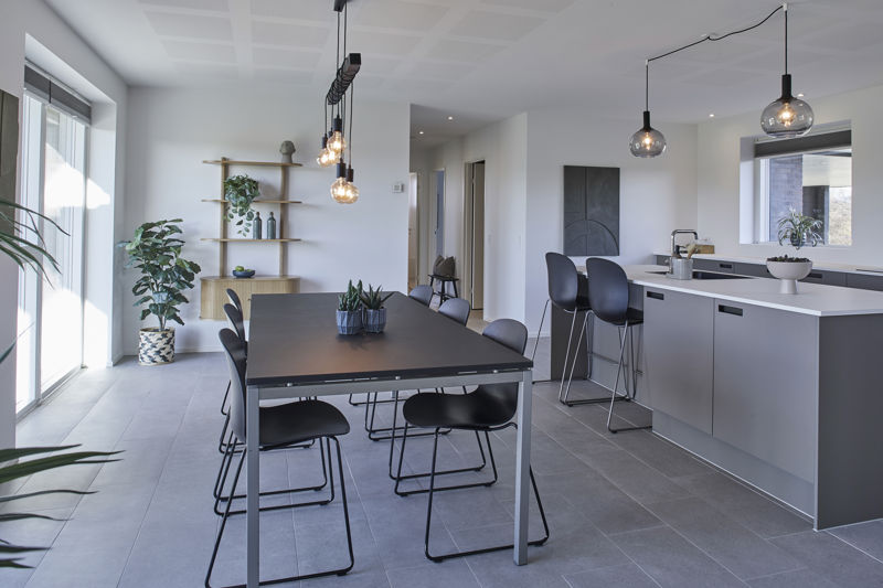 Køkken-alrum fra eurodan-huse med sort spisebord, sorte stole, grå køkkenø, to barstole, sort armatur, runde røgfarvede pendler, åben træhylde med dekoration og stor plante ved vinduet – inspiration til moderne nybyggeri, husbyggeri og køkken-alrum med tydelig kontrast.