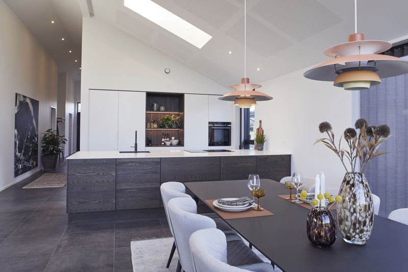 Køkken-alrum fra eurodan-huse med mørk køkkenø, sort armatur, hvide højskabe, åben niche med træhylder, integreret ovn, dækket spisebord i forgrunden, store farvede pendler og lyst loft med ovenlys – inspiration til nybyggeri, husbyggeri og gennemtænkt køkkenindretning.