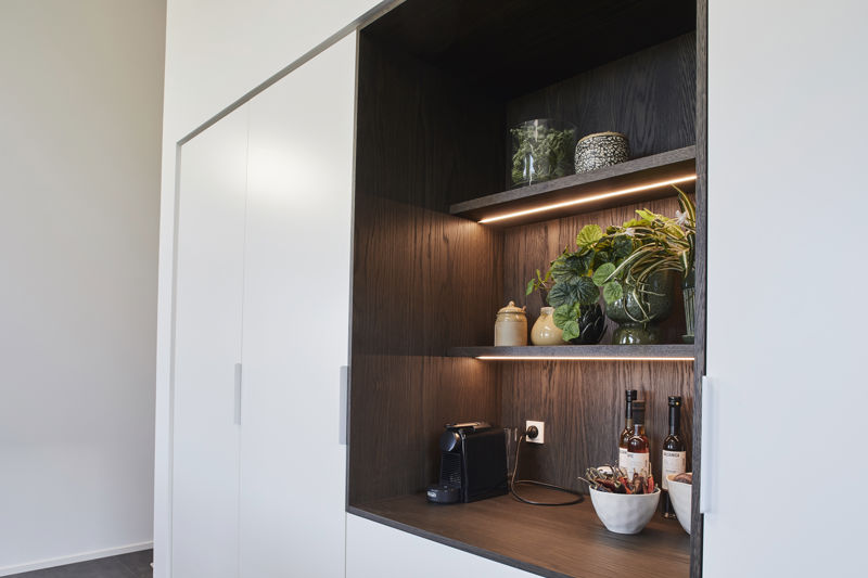 Detalje af indbygget kaffebar og opbevaringsniche fra eurodan-huse med mørkt træ, åbne hylder, integreret LED-lys, grønne planter, kaffemaskine, vinflasker og hvide højskabe på hver side – inspiration til skjult opbevaring, køkkendetaljer og funktionelt nybyggeri.