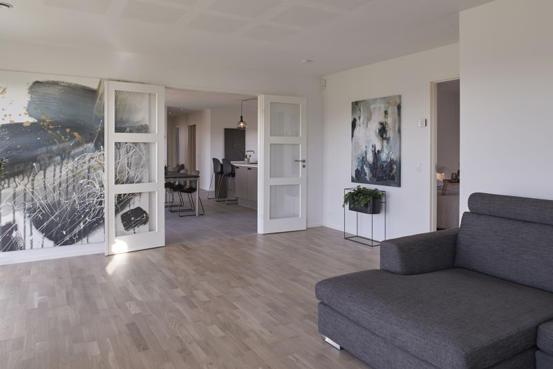 Stue fra eurodan-huse med grå chaiselongsofa, stort abstrakt maleri, dobbeltdøre med matterede felter, åben forbindelse til køkken-alrum, sort plantebord og lyst trælook gulv – konkret inspiration til nybyggeri, husbyggeri og fleksible opholdsrum.