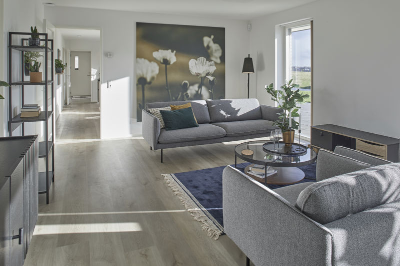 Stue fra eurodan-huse med grå sofa, stor blomsterplakat på væggen, rundt glas-sofabord, mørkeblåt gulvtæppe, sort gulvlampe, lav tv-skænk, sort reol og stort vindue mod terrasse – inspiration til moderne nybyggeri, husbyggeri og indretning af nybygget hus.