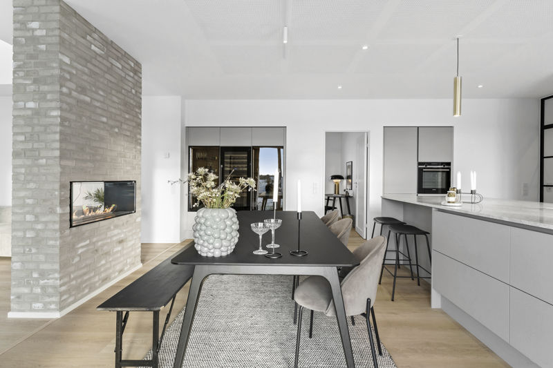 Moderne køkken-alrum fra eurodan-huse med sort spisebord, sort bænk, grå stole, lysegrå køkkenø, barstole, indbygget vinopbevaring, åben barhylde med glas og stor grå murstensvæg med pejseindsats – inspiration til eksklusivt nybyggeri, funktionelt husbyggeri og køkken-alrum med vinløsning.
