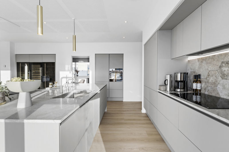 Detalje fra moderne køkken fra eurodan-huse med lang lysegrå køkkenø, dobbelt vask, højt armatur, marmoreret bordplade, pendler i messinglook, kaffestation langs væggen, stænkplade i natursten og integreret ovn i højskab – inspiration til nybyggeri, køkkenfunktioner og moderne husdrømme.