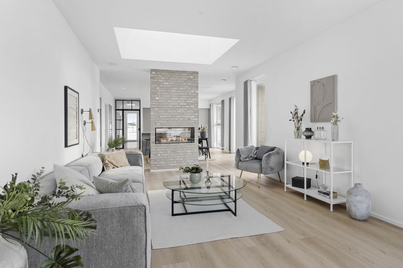 Stue fra eurodan-huse med grå sofa, rund glas-sofabord, hvid reol, grå lænestol, stor murstenspejs som rumdeler, ovenlys og åben forbindelse til spiseplads – inspiration til nybyggeri, husbyggeri og gennemtænkte opholdsrum.