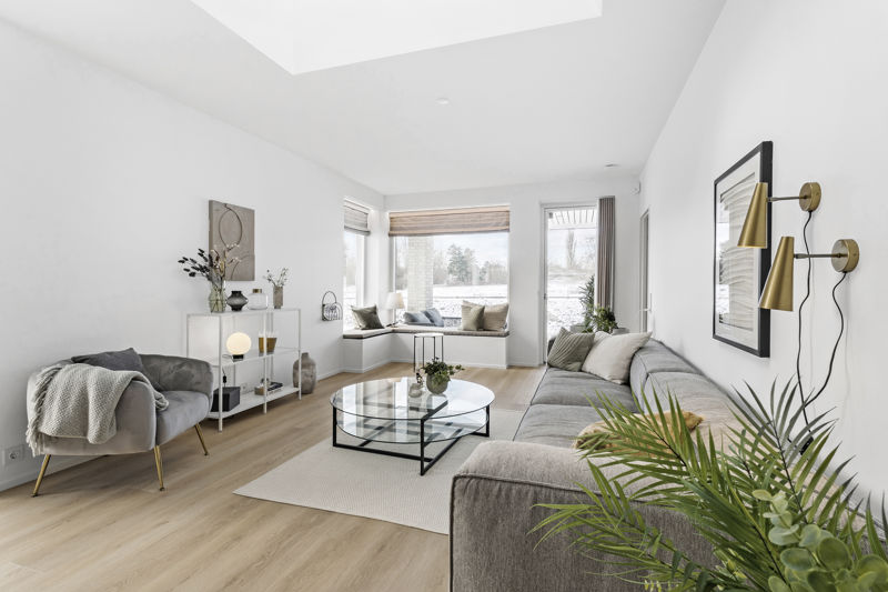Lys stue fra eurodan-huse med grå sofa, rund glas-sofabord, hvid reol med dekoration, grå loungestol, væglamper i messing, sansevindue med hynder og ovenlys – inspiration til nybyggeri, husbyggeri og moderne husdrømme.