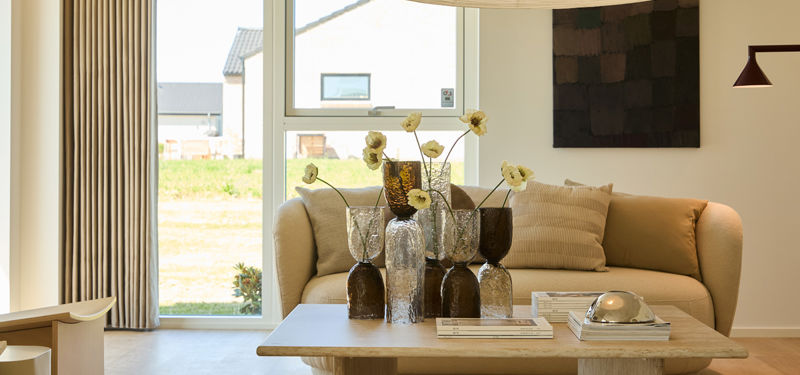 Stue fra eurodan-huse med beige sofa, sofabord med dekorative vaser, stort vindue, gardiner og varm lampebelysning – inspiration til nybyggeri, boligindretning og afslappede husdrømme.