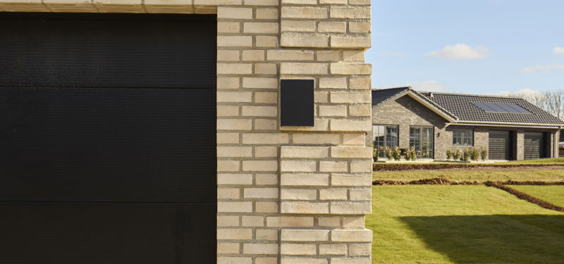 Detalje af husbyggeri med lys murstensfacade, sort garageport og sort tegltag