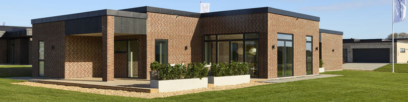 Facade og terrasse fra eurodan-huse med rødbrune mursten, sorte vinduespartier, mørk stern, lavt plantebed foran glasdøre og stor grøn have omkring huset – inspiration til nybyggeri med terrasse, uderum og moderne husdrømme.