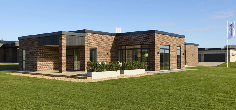 Facade og terrasse fra eurodan-huse med rødbrune mursten, sorte vinduespartier, mørk stern, lavt plantebed foran glasdøre og stor grøn have omkring huset – inspiration til nybyggeri med terrasse, uderum og moderne husdrømme.