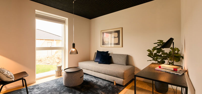 Gæsteværelse fra eurodan-huse med grå sofa, mørkt gulvtæppe, rundt pufbord, sort skrivebord, bordlampe, grøn plante og stort vindue mod haven – inspiration til fleksible rum i nybyggeri og gennemtænkt husbyggeri.