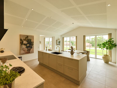 Lyst køkken-alrum fra eurodan-huse med beige køkkenø, messingarmatur, lyse fliser, akustikloft, sprossede vinduer og kig ud til haven – inspiration til moderne nybyggeri.