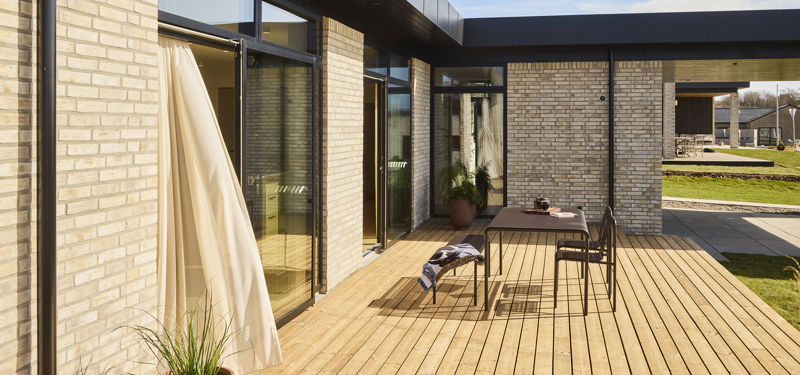 Terrasse fra eurodan-huse med træbelægning, udendørs spiseplads, sort tagudhæng, lyse gardiner ved glasdør, krukkeplanter og delvist overdækket uderum mellem murstensfacader – inspiration til funktionelt nybyggeri, husbyggeri og terrasse i nye huse.