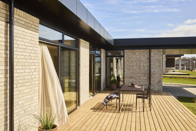 Terrasse fra eurodan-huse med træbelægning, udendørs spiseplads, sort tagudhæng, lyse gardiner ved glasdør, krukkeplanter og delvist overdækket uderum mellem murstensfacader – inspiration til funktionelt nybyggeri, husbyggeri og terrasse i nye huse.