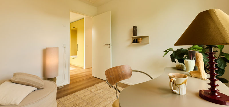 Kontor fra eurodan-huse med rundt skrivebord, designstol, bordlampe, åben dør til gang og kig mod gul skabsvæg – inspiration til fleksibelt nybyggeri.
