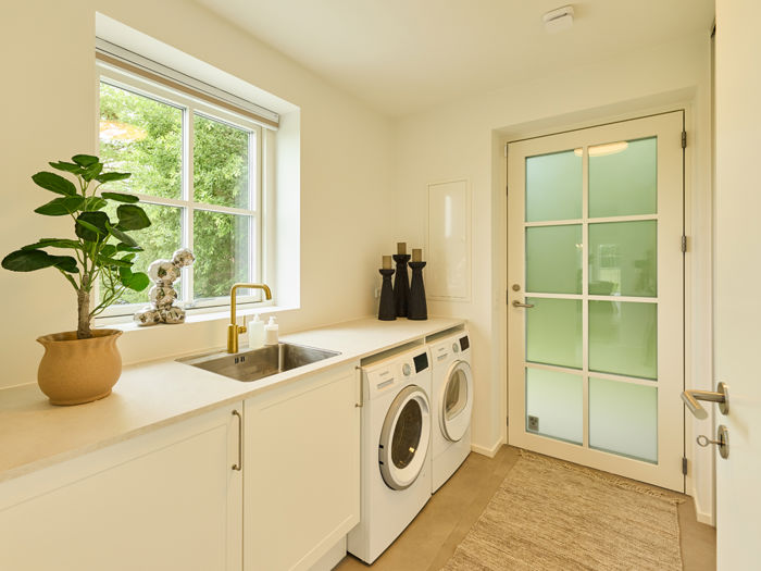 Bryggers fra eurodan-huse med hvid bordplade, vaskemaskine og tørretumbler under bordet, lille stålvask, messingarmatur, vindue og dør med matteret glas – inspiration til praktisk nybyggeri.
