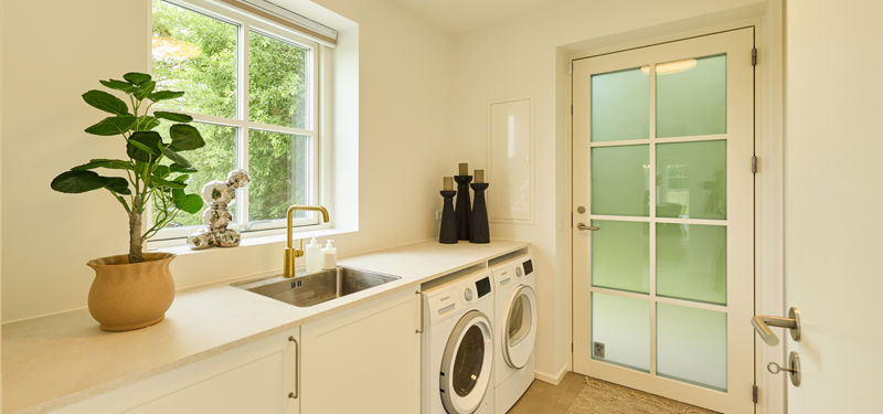 Bryggers fra eurodan-huse med hvid bordplade, vaskemaskine og tørretumbler under bordet, lille stålvask, messingarmatur, vindue og dør med matteret glas – inspiration til praktisk nybyggeri.