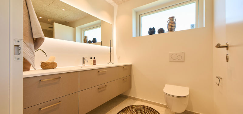Badeværelse fra eurodan-huse med langt vaskemøbel, stort spejl med indirekte lys, væghængt toilet, højtsiddende vindue og beige gulvfliser – inspiration til funktionelt nybyggeri og husdrømme.