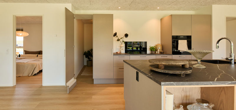 Køkken fra eurodan-huse med beige skabsvæg, integreret ovn, mørk bordplade, stor køkkenø og åben låge ind til depot/opbevaring – inspiration til funktionelt nybyggeri og gennemtænkte husdrømme.