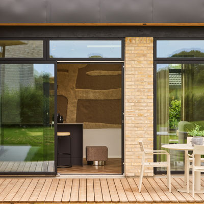 Kig fra terrasse ind mod køkken-alrum fra eurodan-huse med sorte skydedøre, høje glaspartier, lys murstensfacade, terrassebord, havemøbler og direkte forbindelse mellem udeområde og køkken – inspiration til nybyggeri med terrasse og husdrømme.