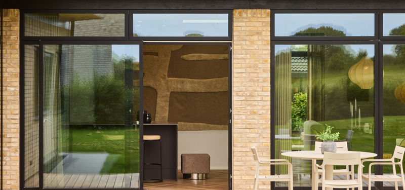 Kig fra terrasse ind mod køkken-alrum fra eurodan-huse med sorte skydedøre, høje glaspartier, lys murstensfacade, terrassebord, havemøbler og direkte forbindelse mellem udeområde og køkken – inspiration til nybyggeri med terrasse og husdrømme.