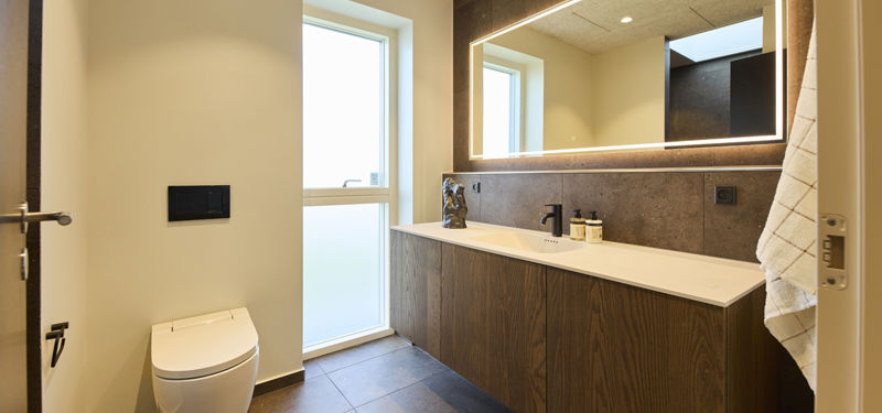 Badeværelse fra eurodan-huse med væghængt toilet, sort betjeningsplade, højt vindue, langt træmøbel, sort armatur, mørk flisevæg bag vasken og stort spejl med lysramme – inspiration til nybyggeri med moderne badeværelsesindretning.