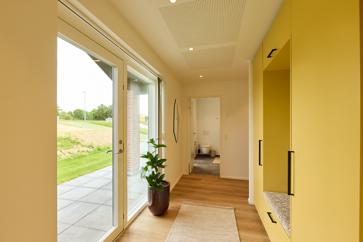 Entré fra eurodan-huse med glasdør, stort sidevindue, gul skabsvæg, indbygget siddeniche, plante og kig til badeværelse – inspiration til praktisk nybyggeri.