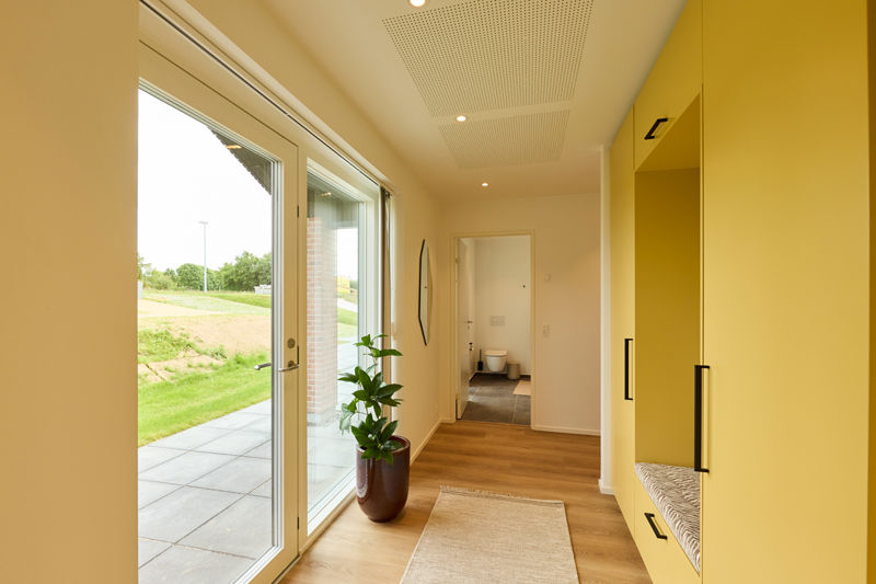 Entré fra eurodan-huse med glasdør, stort sidevindue, gul skabsvæg, indbygget siddeniche, plante og kig til badeværelse – inspiration til praktisk nybyggeri.