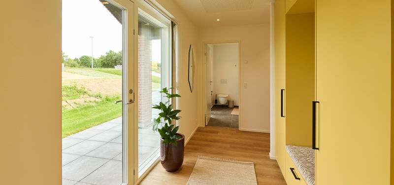 Entré fra eurodan-huse med glasdør, stort sidevindue, gul skabsvæg, indbygget siddeniche, plante og kig til badeværelse – inspiration til praktisk nybyggeri.