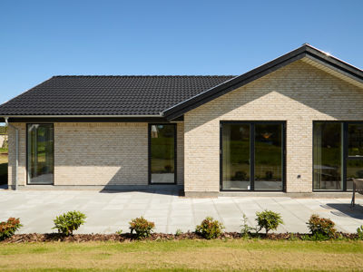 Facade fra eurodan-huse med lyse mursten, mørkt tag, sorte vinduespartier, flisebelagt terrasse og beplantning foran huset – inspiration til nybyggeri, husbyggeri og moderne husdrømme.