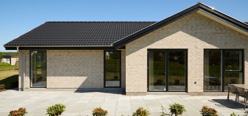 Facade fra eurodan-huse med lyse mursten, mørkt tag, sorte vinduespartier, flisebelagt terrasse og beplantning foran huset – inspiration til nybyggeri, husbyggeri og moderne husdrømme.