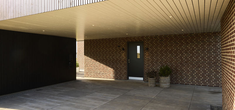 Overdækket carport ved moderne vinkelhus med mørk træbeklædning, indbyggede spots og indgangsdør – inspiration til nyt hus.