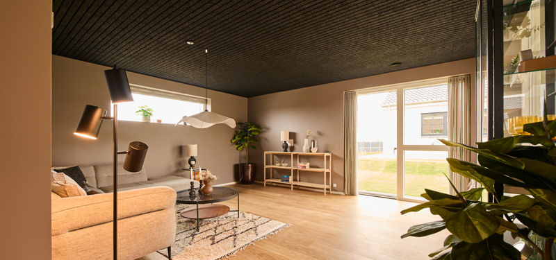 Lys opholdsstue med sofaområde, mørkt loft og store vinduespartier med udsigt til haven – en hyggelig ramme om familielivet i drømmehuset.