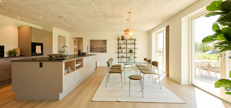 Køkken-alrum fra eurodan-huse i hele rummets bredde med beige køkkenø, åbne hylder, glasbord, spisebordsstole, store vinduer og terrasseudgang – inspiration til moderne nybyggeri og husdrømme.