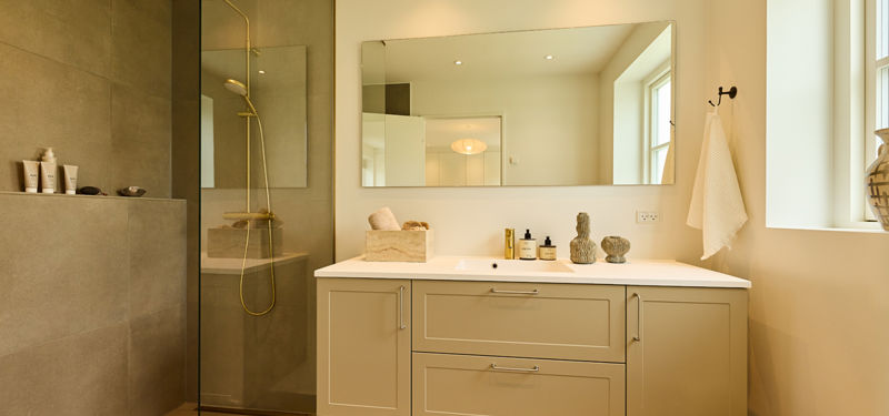 Badeværelse fra eurodan-huse med beige badmøbel, stor spejlvæg, messingarmatur, bruseniche med glasvæg, grå fliser og smalt vindue – inspiration til moderne nybyggeri.