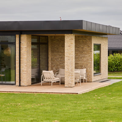 Overdækket træterrasse fra eurodan-huse med lyse murstenssøjler, sort udhæng, store glaspartier, loungestole og tæt forbindelse til boligens opholdsrum – inspiration til nybygget hus med overdækket uderum og moderne husbyggeri.
