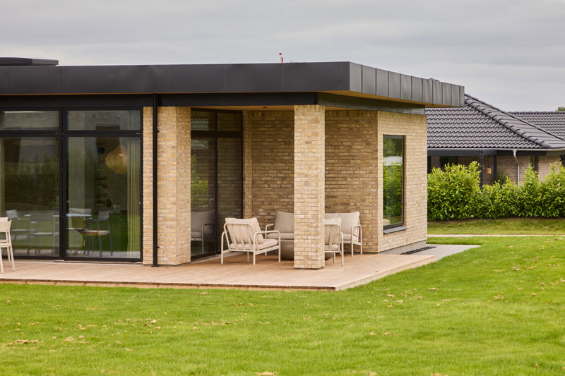Overdækket træterrasse fra eurodan-huse med lyse murstenssøjler, sort udhæng, store glaspartier, loungestole og tæt forbindelse til boligens opholdsrum – inspiration til nybygget hus med overdækket uderum og moderne husbyggeri.