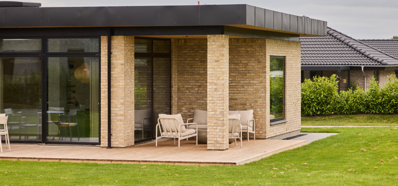Overdækket træterrasse fra eurodan-huse med lyse murstenssøjler, sort udhæng, store glaspartier, loungestole og tæt forbindelse til boligens opholdsrum – inspiration til nybygget hus med overdækket uderum og moderne husbyggeri.