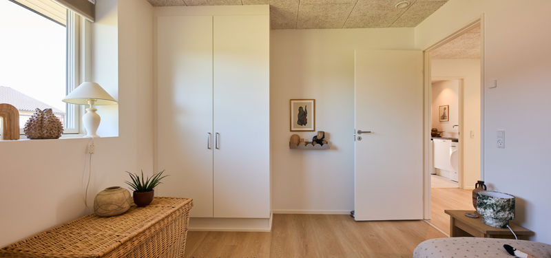 Værelse fra eurodan-huse med hvidt indbygget skab, lavt vindue, fletopbevaring, plante, åben dør og kig mod badeværelse – inspiration til praktisk indretning, nybyggeri og husdrømme.