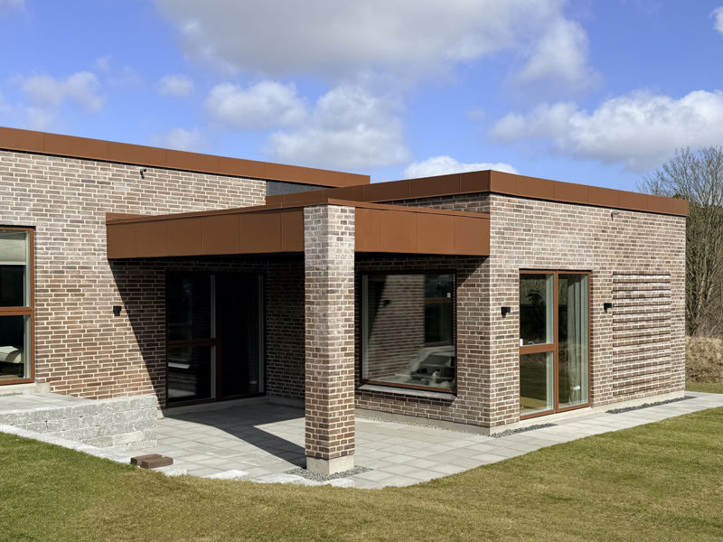 Overdækket terrasse fra eurodan-huse med grå terrassefliser, lyse mursten, brune vinduesrammer, fladt tagudhæng og søjler, der skaber læ ved facaden – inspiration til moderne terrasse, nybyggeri og arkitektoniske husdrømme.