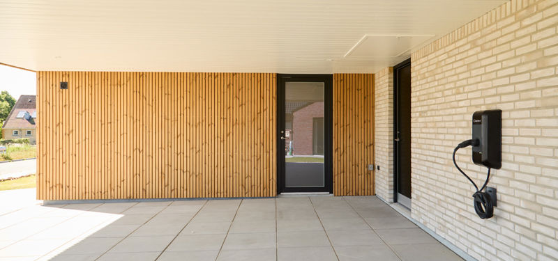 Carport fra eurodan-huse med træbeklædt væg, lys murstensfacade, sort dør, flisebelægning og el-lader på væggen – inspiration til nybyggeri med carport, praktisk hverdag og husdrømme.