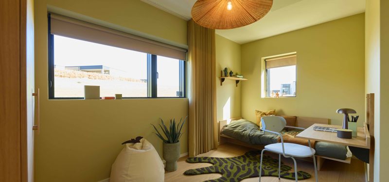 Børneværelse fra eurodan-huse med grønne vægge, seng i hjørnet, skrivebord, hvid stol, bredt vindue, beige gardiner, grøn mønstret løber og stor flettet loftlampe – inspiration til børneværelser, nybyggeri og husdrømme.