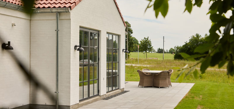 Terrasse ved hvid facade fra eurodan-huse med rødt tegltag, sorte sprossede terrassedøre, udendørslamper, loungemøbler og flisebelægning – inspiration til uderum ved nybyggeri.